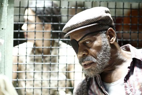 Danny Glover seated behind a cage-like gate as if in a prison. He is wearing one eyepatch on his right eye and his left eye is cloudy. He has a scruffy beard and is wearing a newsboy cap. The focus is on him and you can see someone behind the screen.